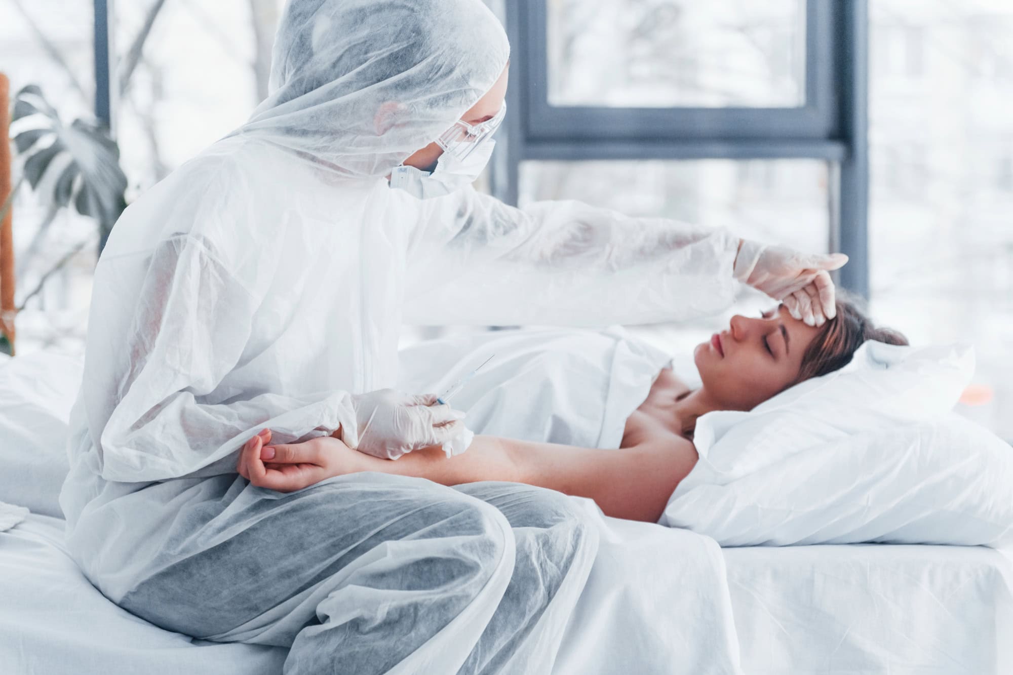 Doctor in PPE checking forehead of a female patient lying in bed