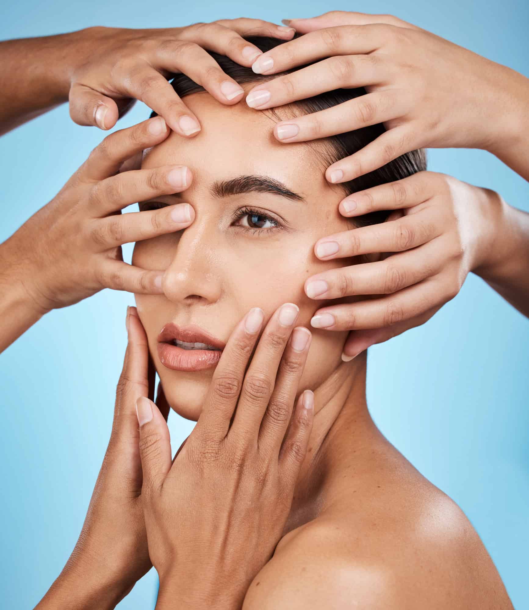 Close-up portrait of a woman surrounded by multiple hands touching her face against a blue background.