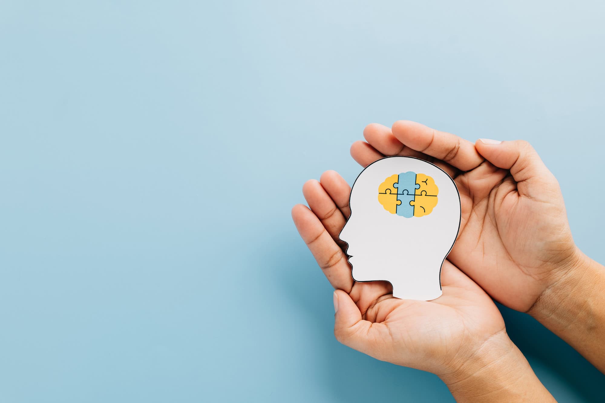 Hands holding a paper cutout of a human head with a brain made of puzzle pieces on a blue background.